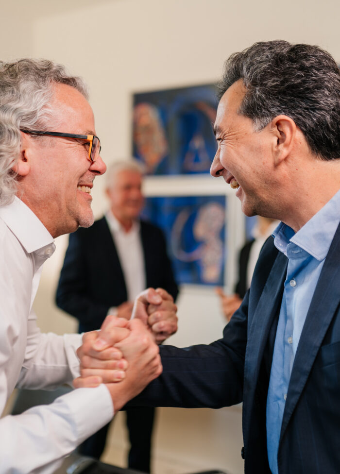 Two men are smiling and shaking hands enthusiastically, with a blurred figure in the background and abstract artwork on the walls.