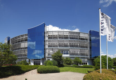 Ein modernes Firmengebäude mit reflektierenden Fensterfronten und grünen Büschen im Vordergrund, unter blauem Himmel.