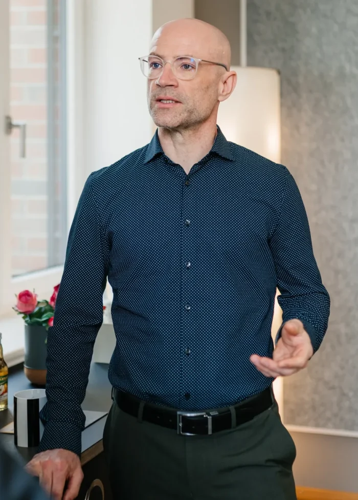 Zwei Personen stehen in einem modern eingerichteten Raum, während einer von ihnen in einem blauen Hemd mit Muster spricht und gestikuliert, während der andere in einem dunklen Anzug ruhig zuhört.