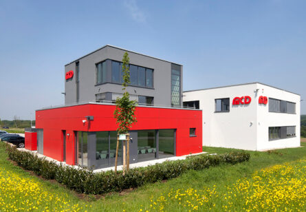 Zwei moderne Bürogebäude mit roten und grauen Fassaden, umgeben von grünen Wiesen und gelben Blumen, stehen unter einem blauen Himmel.